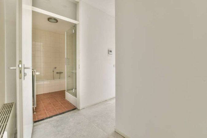 Couloir étroit aux murs blancs donnant sur une salle de bain carrelée avec cabine de douche en verre et baignoire, dans un appartement épuré.