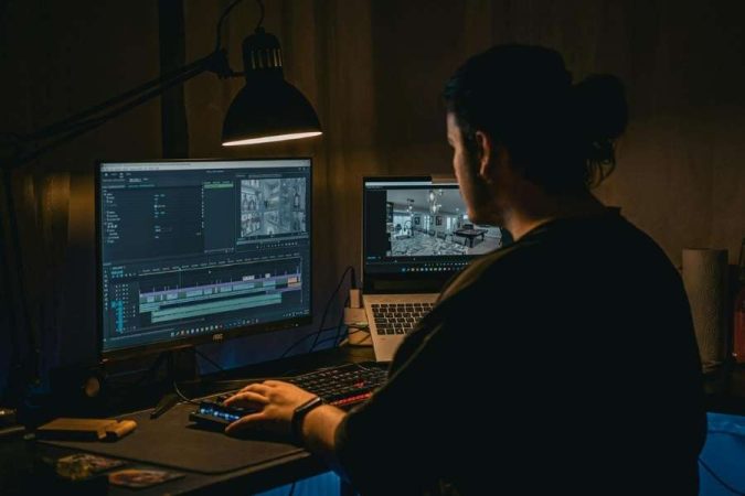 Un monteur vidéo de dos travaille sur son ordinateur de bureau et son ordinateur portable dans une pièce sombre éclairée par une lampe de bureau.