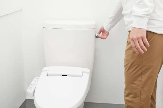 Un homme debout actionne la chasse d'eau de ses toilettes modernes dans une pièce aux murs blancs.